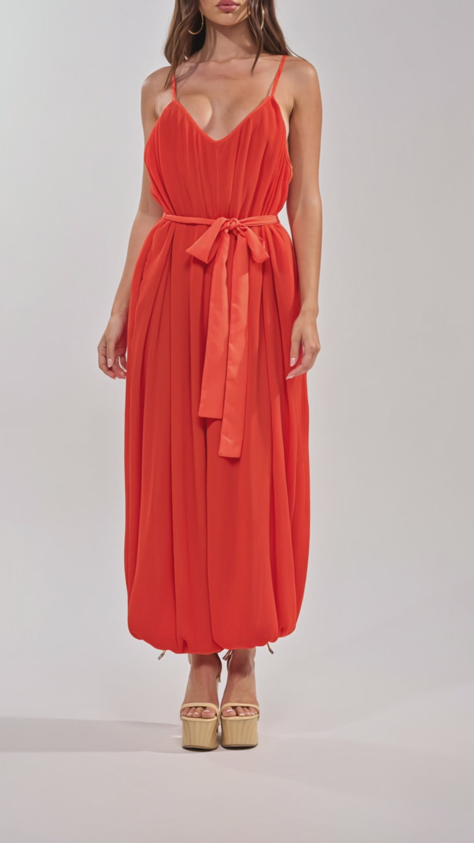 Video of woman modeling the VACATIONING PLEATED JUMPSUIT IN ORANGE with a tied waist and ankle cuffs, paired with beige platform heels.