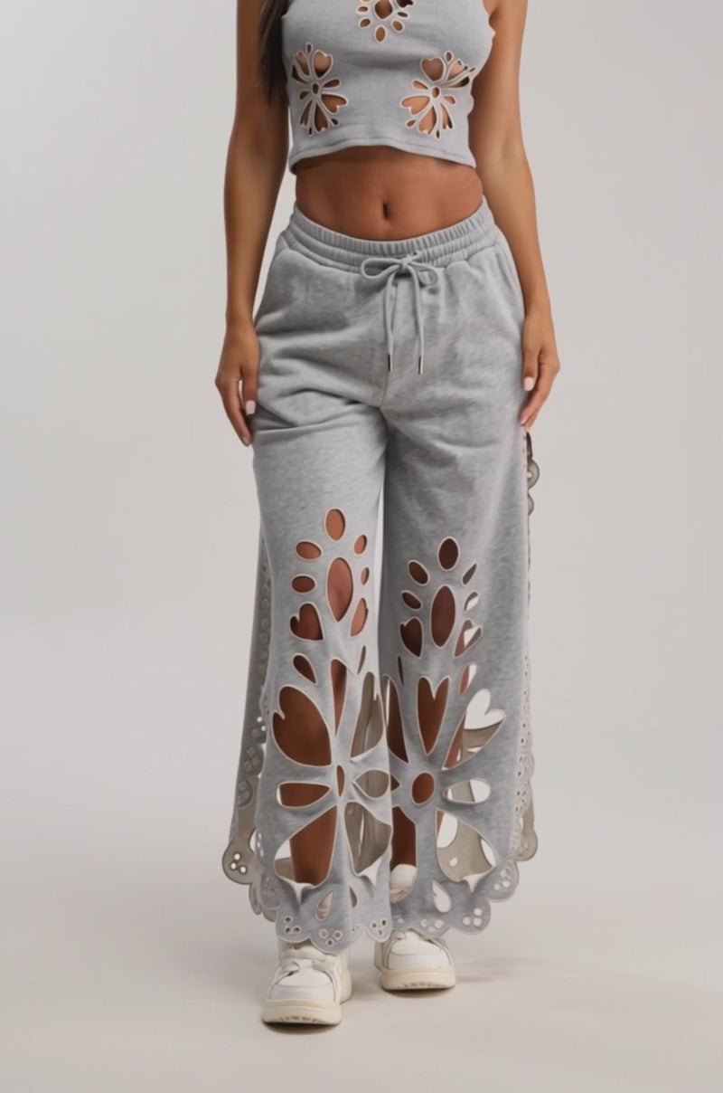 Video of woman modeling the GIRLY POP CUTOUT JOGGER set in heather grey with floral cutouts, wide-leg pants, and white sneakers, posing with one hand in her pocket against a plain background.