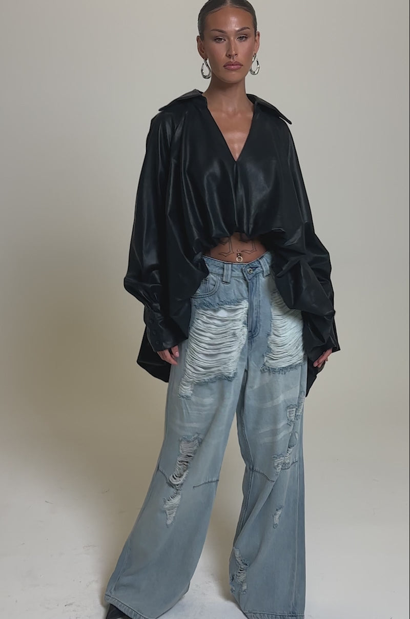Video of woman modeling the NEW ATTITUDE FAUX LEATHER HI-LOW BLOUSE with voluminous sleeves, styled with light blue ripped jeans, slicked-back hair, hoop earrings, and a navel piercing.