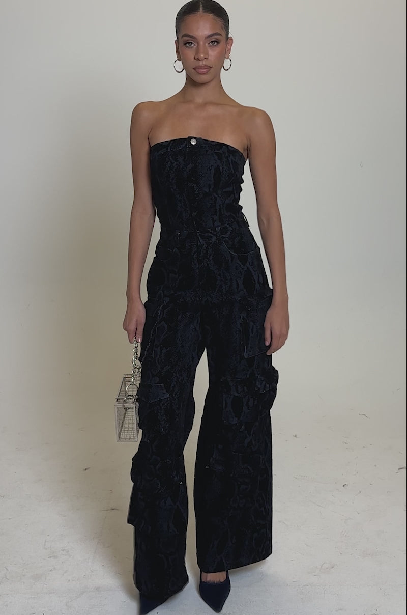 A video of a woman posing against a plain backdrop in the CAUGHT YOUR EYE VELVET DENIM JUMPSUIT IN SNAKEPRINT, featuring wide legs. She accessorizes with a metallic handbag, hoop earrings, and dark heels, wearing her hair pulled back for a stylish touch.