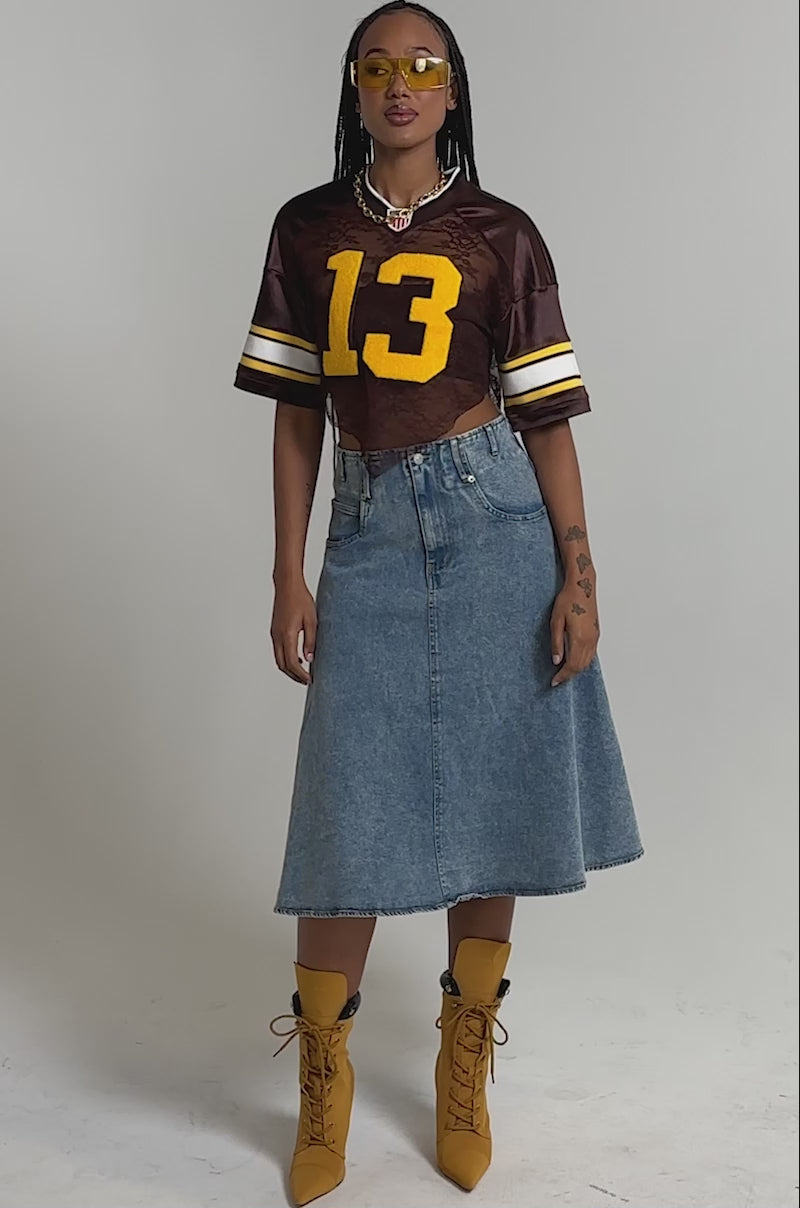 A video of a woman in the LUCKY LACE JERSEY TOP IN BROWN with the number 13 and varsity patch, paired with a blue denim midi skirt, mustard lace-up heeled boots, and yellow sunglasses.
