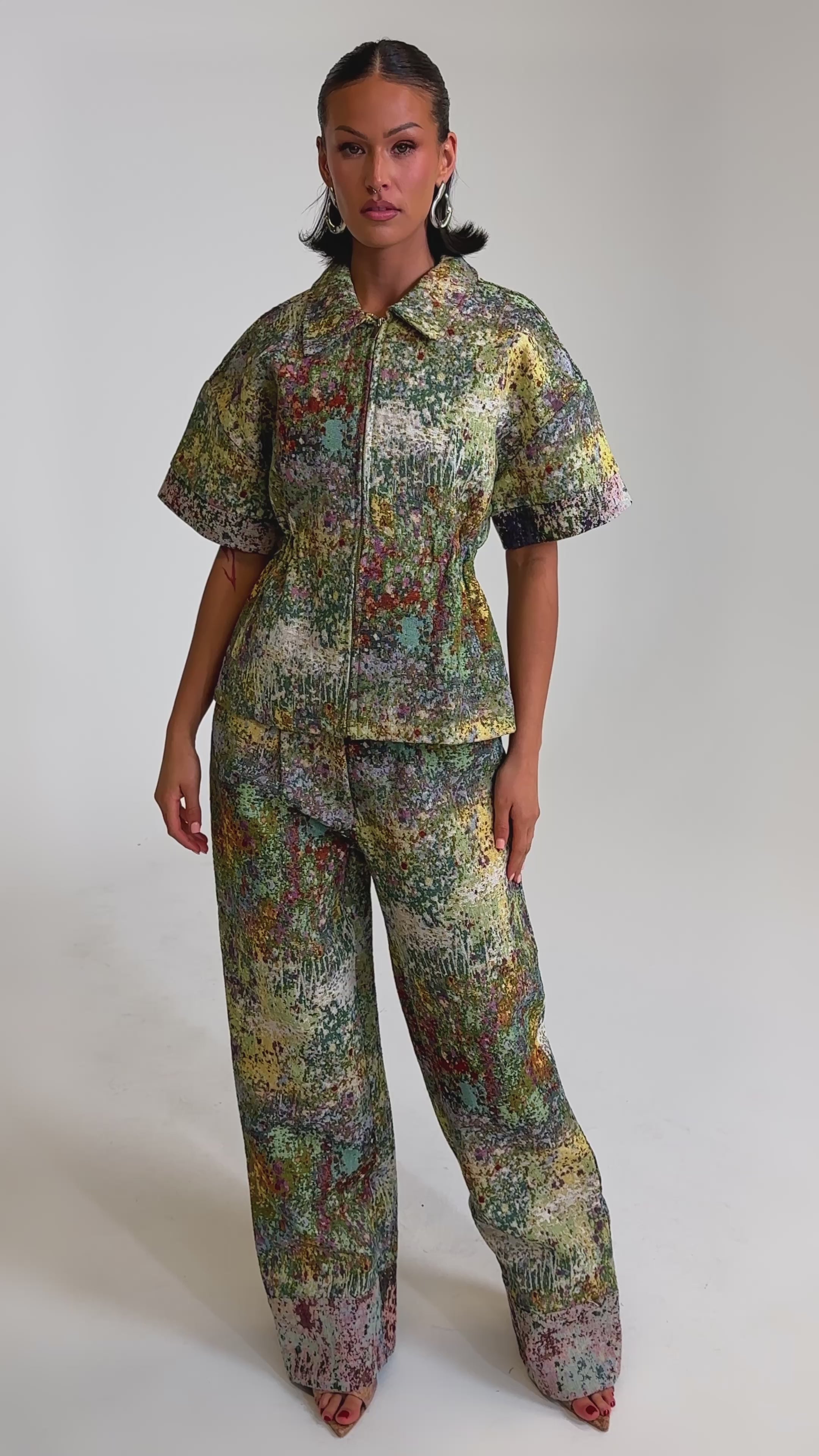 A video of a woman in DEEP IN THE SEA BROCADE TROUSER—a high-rise, wide-leg pant with an abstract, multicolored pattern—paired with a matching loose short-sleeve button-up shirt against a plain light background.