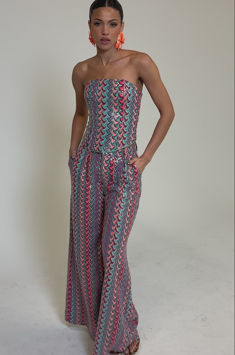 Video of woman modeling the COASTAL BABE TROUSER, a colorful, strapless wide-leg pant with a blue, pink, and yellow wavy pattern—ideal for vacations. She completes the look with bright orange earrings and slicked-back hair.