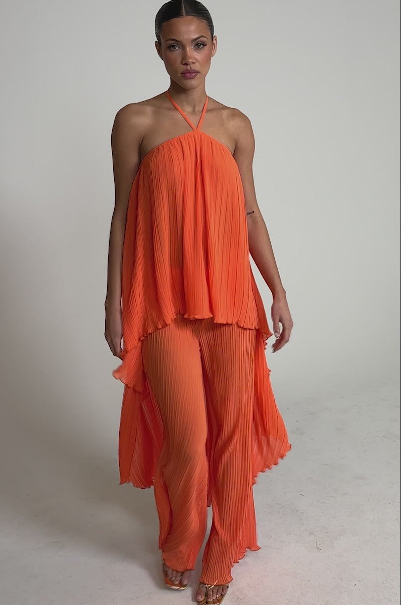 Video of woman modeling the CRUSHIN ON YOU PALAZZO PANT, featuring bright orange pleats and an asymmetrical halter-neck top—ideal for adding flair to your vacation look. She pairs it with large hoops and strappy heels against a light backdrop.