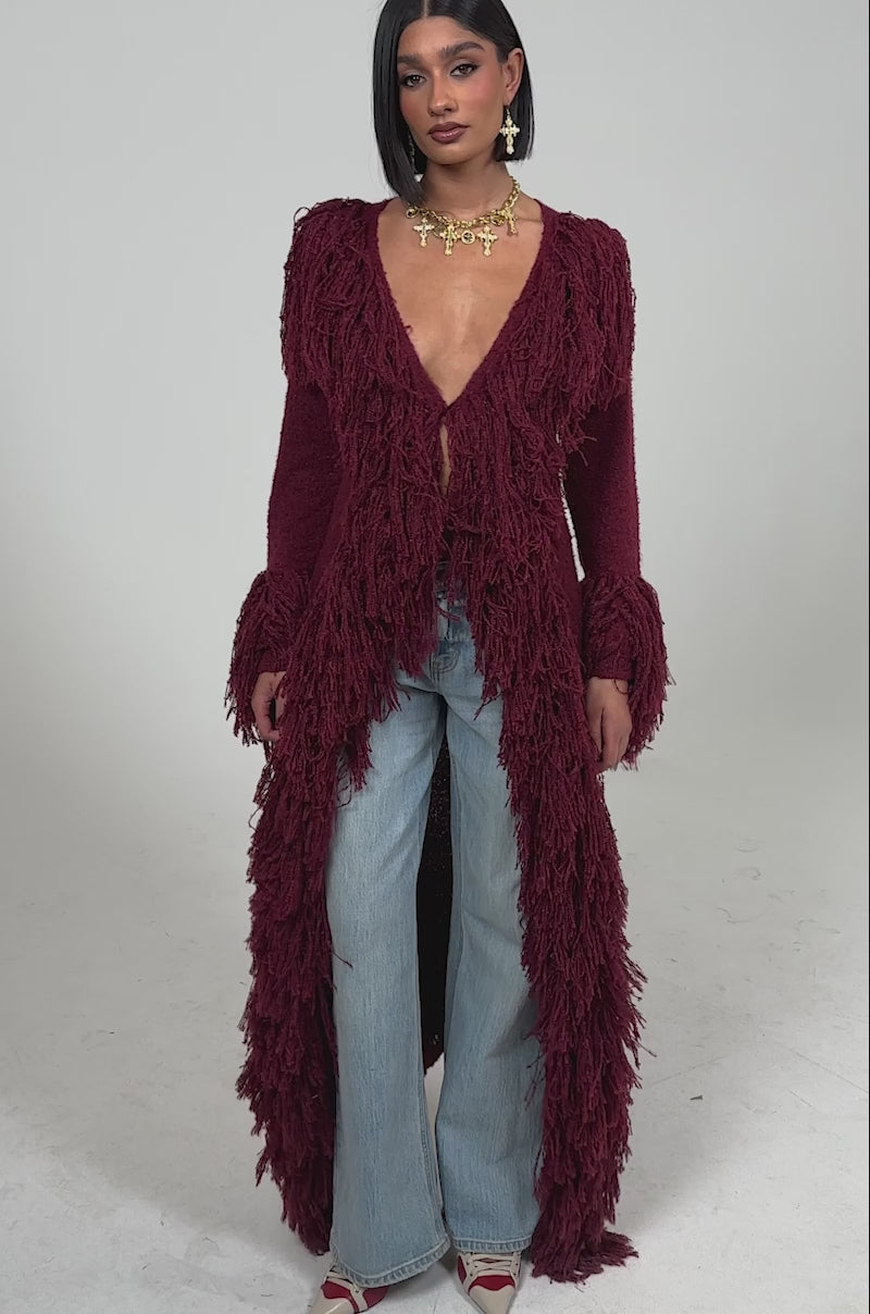 Video of woman modeling the CAN'T GET ENOUGH FRINGE CARDIGAN, styled with a plunging neckline, light-wash flared jeans, pointed heels, and a gold statement necklace against a plain background.