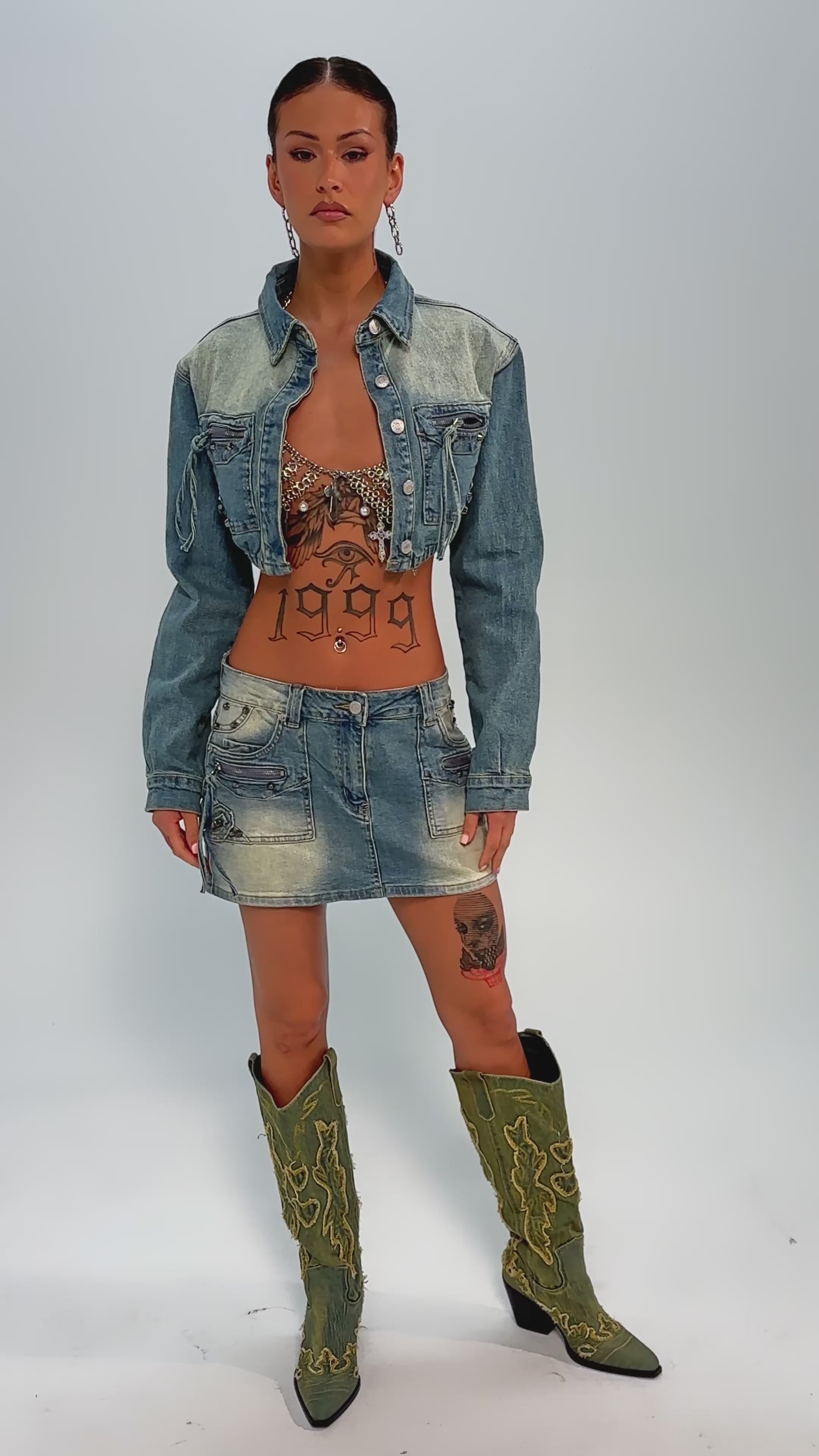 Video of woman modeling the CROWN BELTED DENIM MINI SKIRT and a denim jacket, showing a "1999" tattoo on her stomach and a skull tattoo on her thigh, posed against a neutral background.