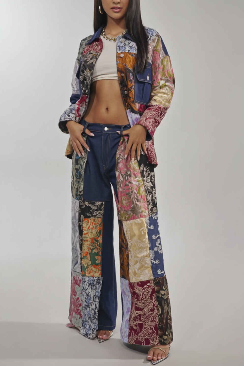 Video of woman modeling the EMOTIONLESS TROUSER wide-leg denim pants with a high-rise waistband, styled with a brocade patchwork denim jacket, white crop top, and necklace against a plain background.