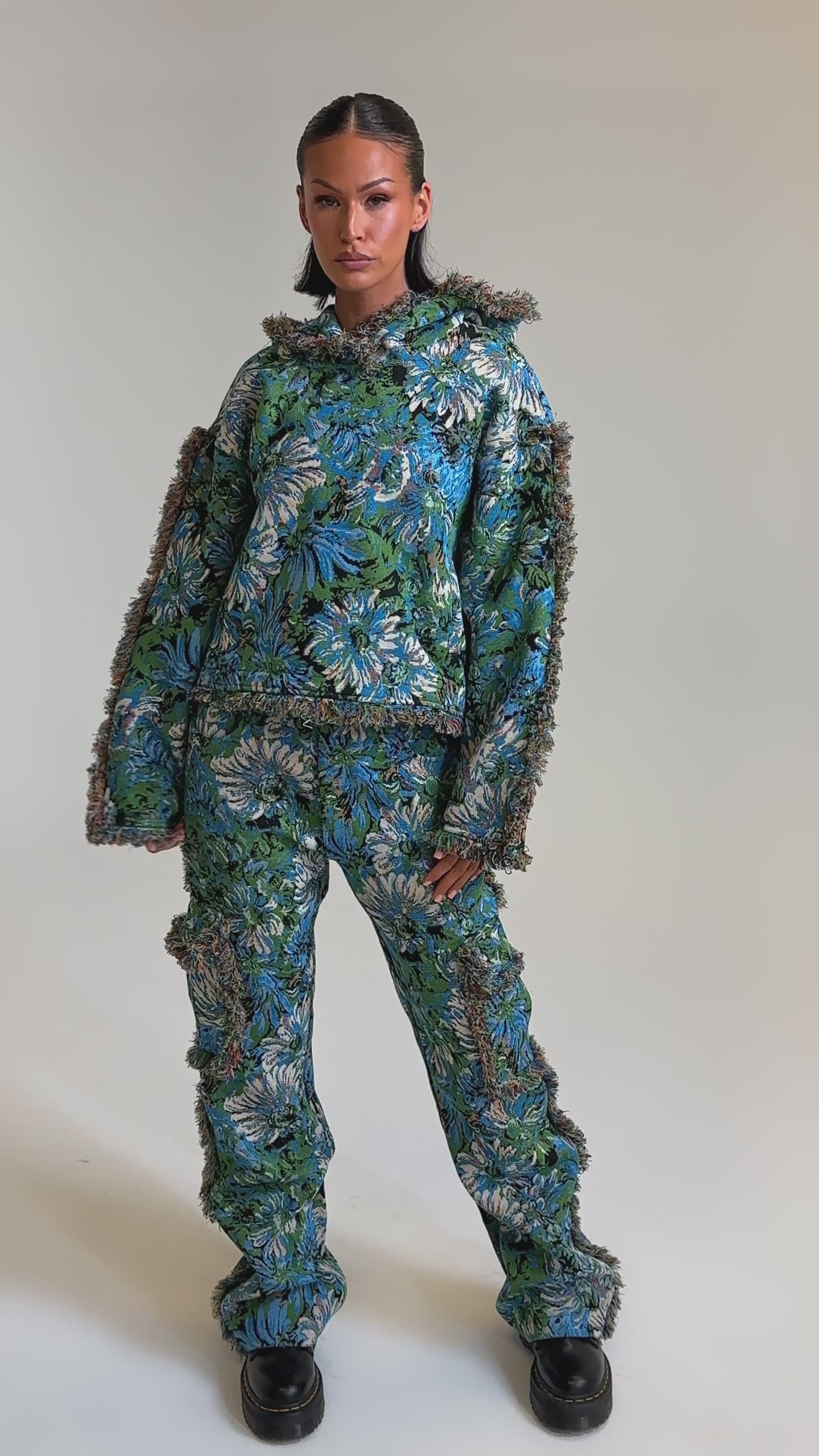 A video of a model wearing the TRY ME TAPESTRY SWEATSHIRT, a person poses in a blue-green floral hooded jacket with fuzzy trim, brown accents, and chunky black shoes—a look that channels vintage sweater charm.