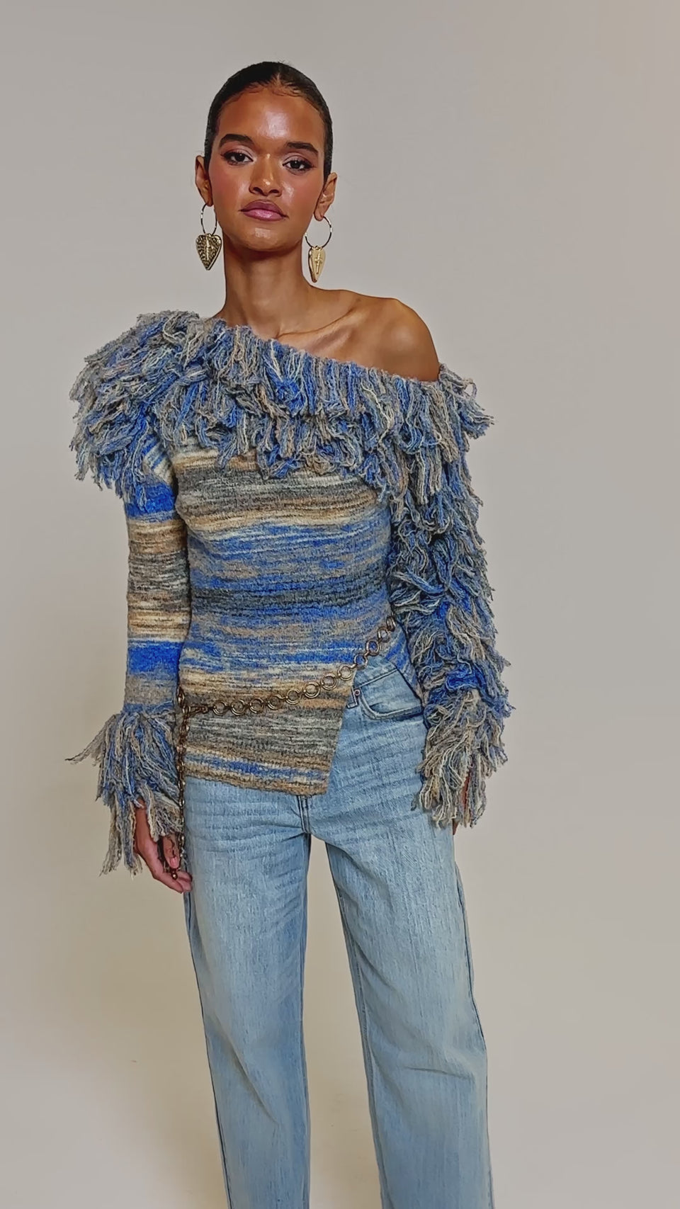 Video of woman modeling the FRINGE FOR DAYS SWEATER—a multicolor striped fringe sweater with one off-the-shoulder sleeve—paired with light blue jeans.