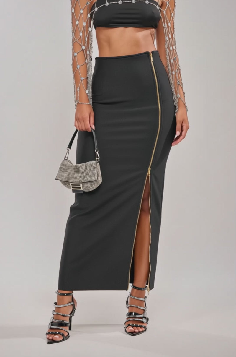 A video of a model wearing the UNZIP ME BANDAGE MAXI SKIRT—a curve-hugging black maxi with a gold zip closure slit—paired with a beaded draped-sleeve top, strappy heeled sandals, and a small silver purse.