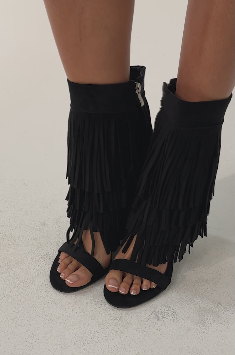 Video of woman modeling the AKIRA GOLD SESTINA BLACK FAUX SUEDE FRINGE WEDGE SANDALS, a black leather mini skirt with a flared hem, and a colorful long-sleeved crochet top. 