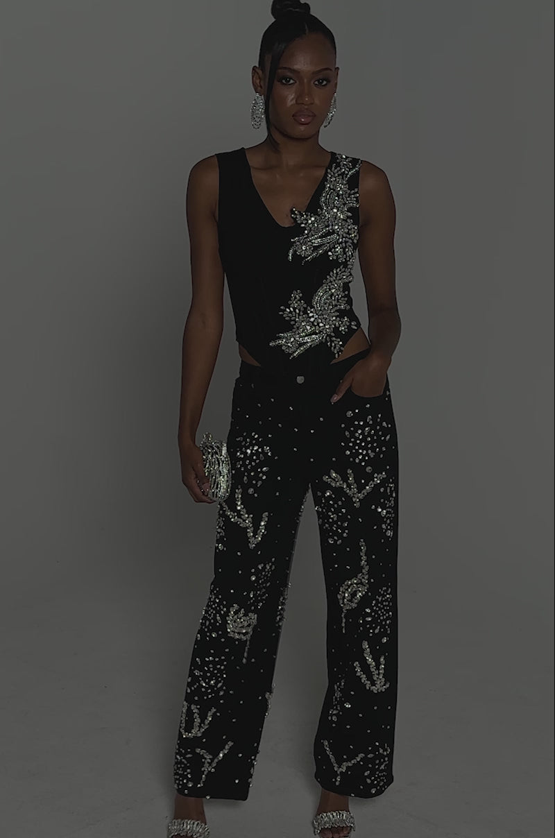 Video of woman modeling the LOTS OF LOVE TO GIVE RHINESTONE BLOUSE, a black sleeveless V-neck top with silver embellishments, paired with matching pants, a silver beaded clutch, and dangling earrings against a plain gray background.