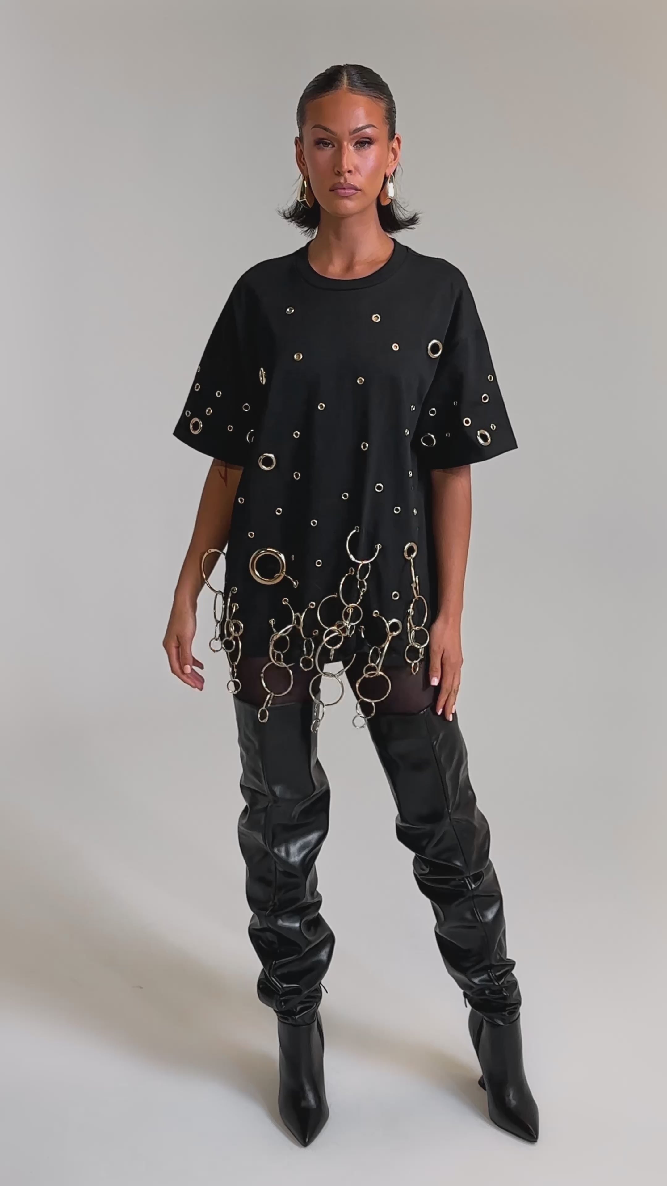 A video of a woman in the RING AROUND ME OVERSIZED TEE, featuring metallic grommets and gold ring details, styled with slouchy black leather thigh-high boots against a plain backdrop for a bold streetwear statement.