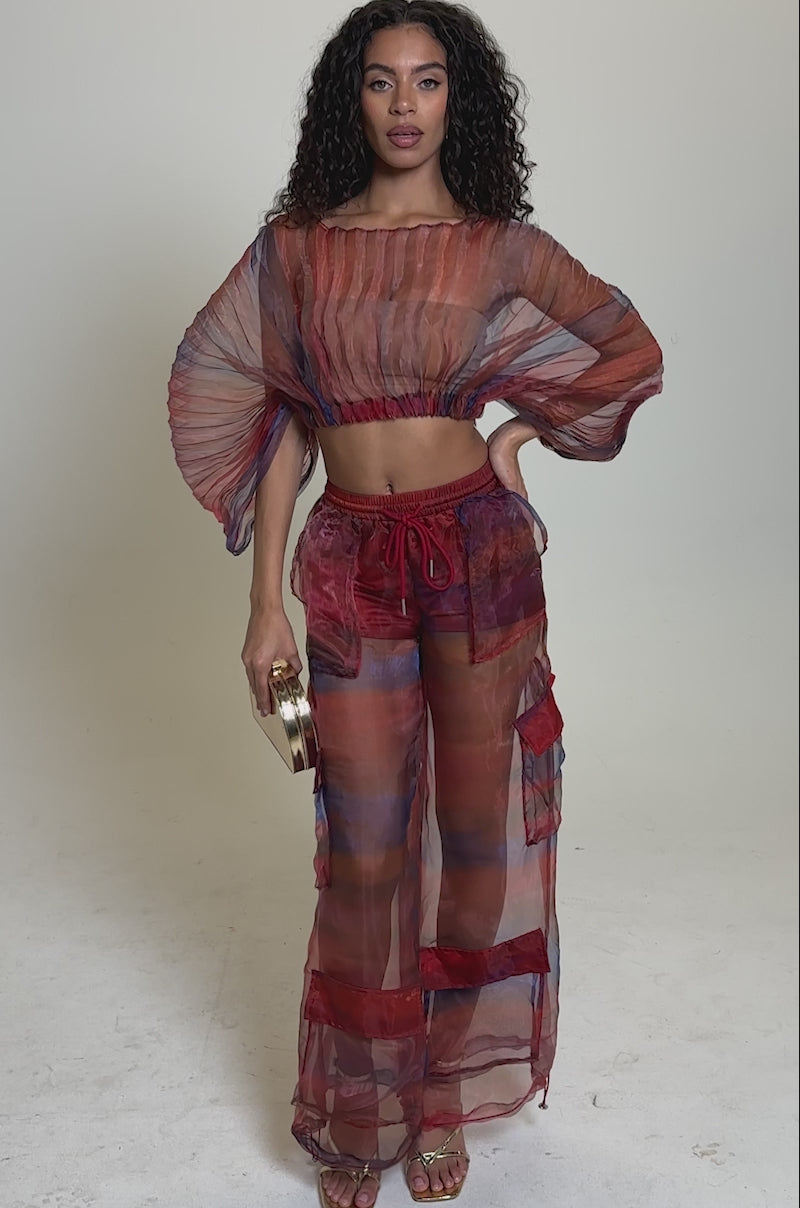 Video of woman modeling the LETS LIVE IN THE MOMENT CHIFFON BLOUSE—a sheer, multicolor organza top paired with matching pants over a red bralette. She holds a gold clutch and faces the camera against a plain background.