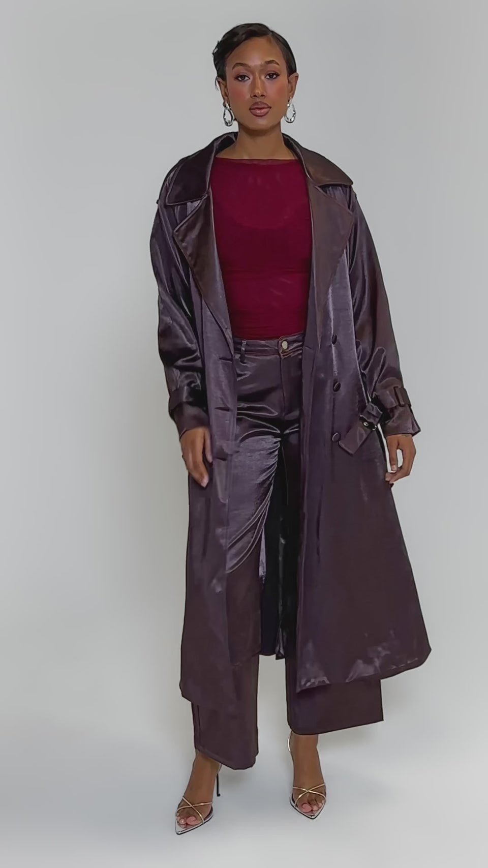 Video of woman modeling the SLOW DOWN SATIN TROUSER, paired with a long mauve satin trench coat, a fitted burgundy top, and strappy high-heeled sandals against a plain light background