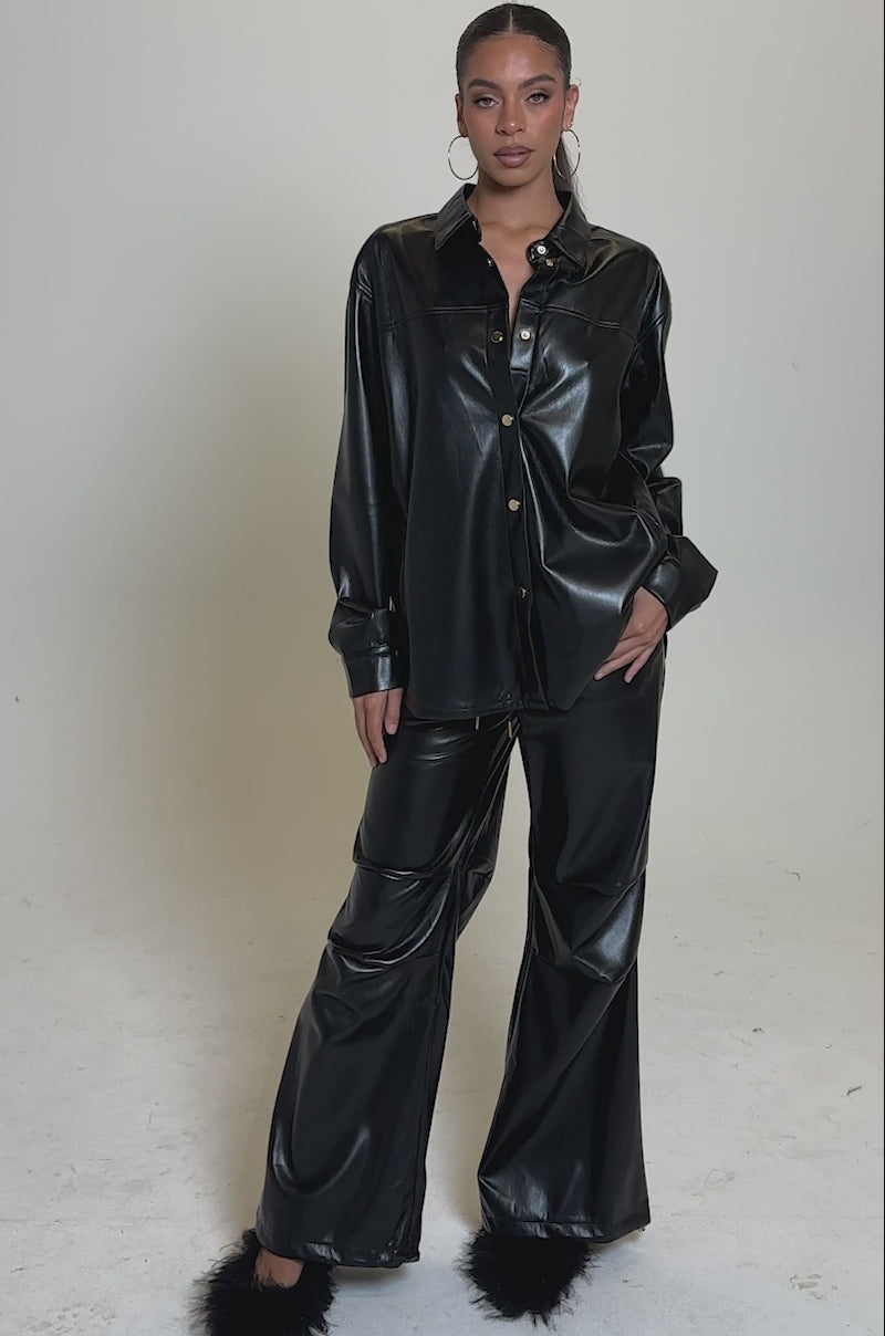 A video of a woman in the GET WITH IT FAUX LEATHER BUTTON DOWN shirt and matching wide-leg pants with black furry footwear, hoop earrings, and her hair pulled back. The plain background is light-colored.