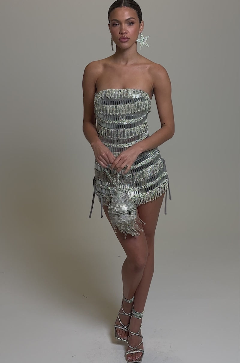 Video of woman modeling the CHANDELIER RHINESTONE MINI DRESS, a strapless metallic stretch piece with reflective details. She holds a sparkling silver bag, wears large star-shaped earrings, and has slicked-back hair against a plain background.