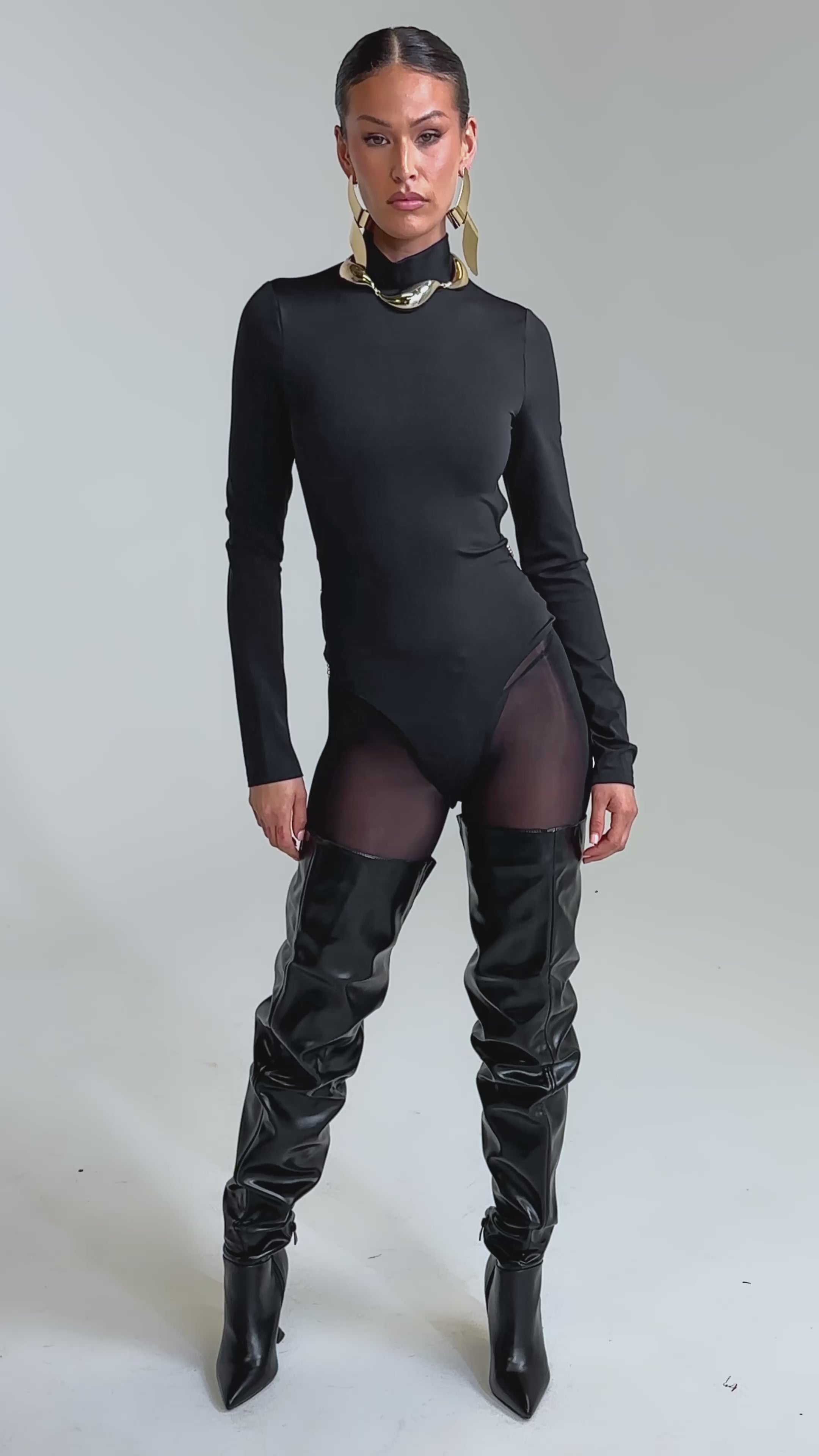 Video of woman modeling the BLACK OUT MOCK NECK OPEN BACK BODYSUIT, featuring sheer fabric and a gold chain belt. She completes the look with large gold earrings.