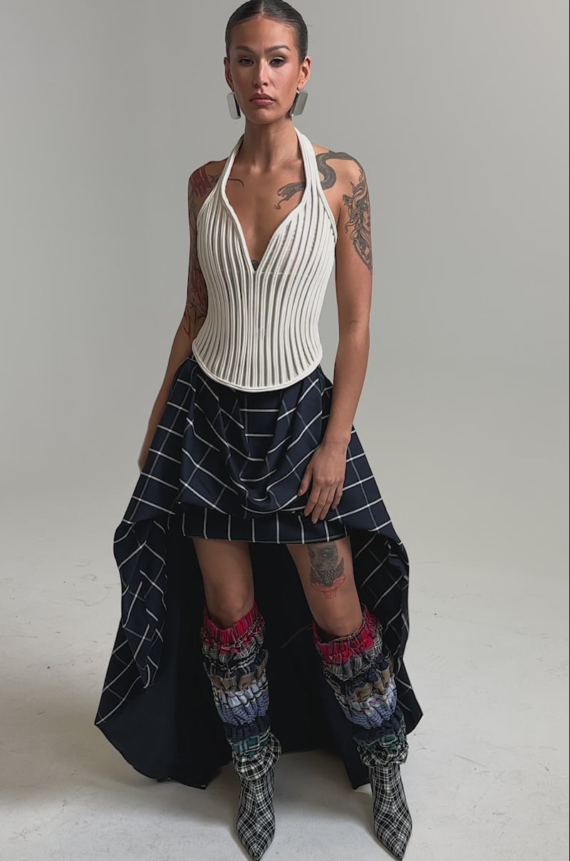 Video of woman modeling the MAKE A STATEMENT HIGH LOW SKIRT, featuring an asymmetrical plaid design with white grid lines, paired with a ribbed cream top, colorful leg warmers, checkered boots, and visible tattoos. Her hair is pulled back.