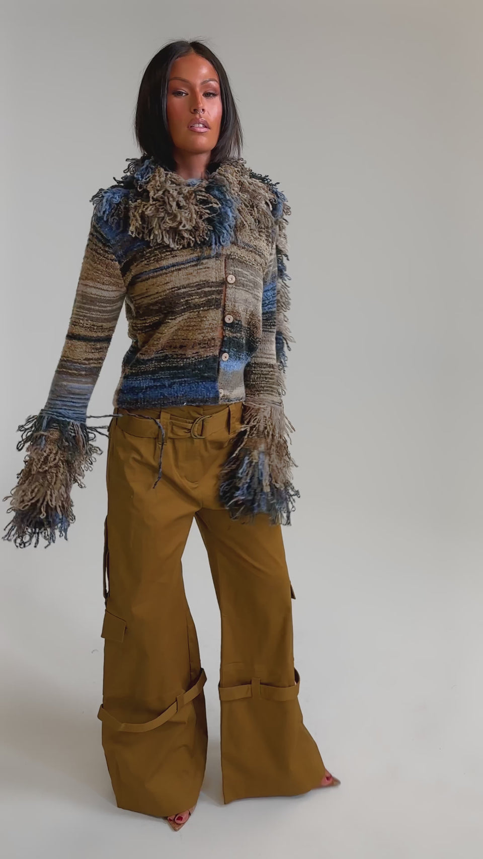 A video of a person with straight dark hair wearing a textured, multicolored sweater with fringe and the DOWN FOR ANYTHING TROUSER, standing confidently against a plain, light background.