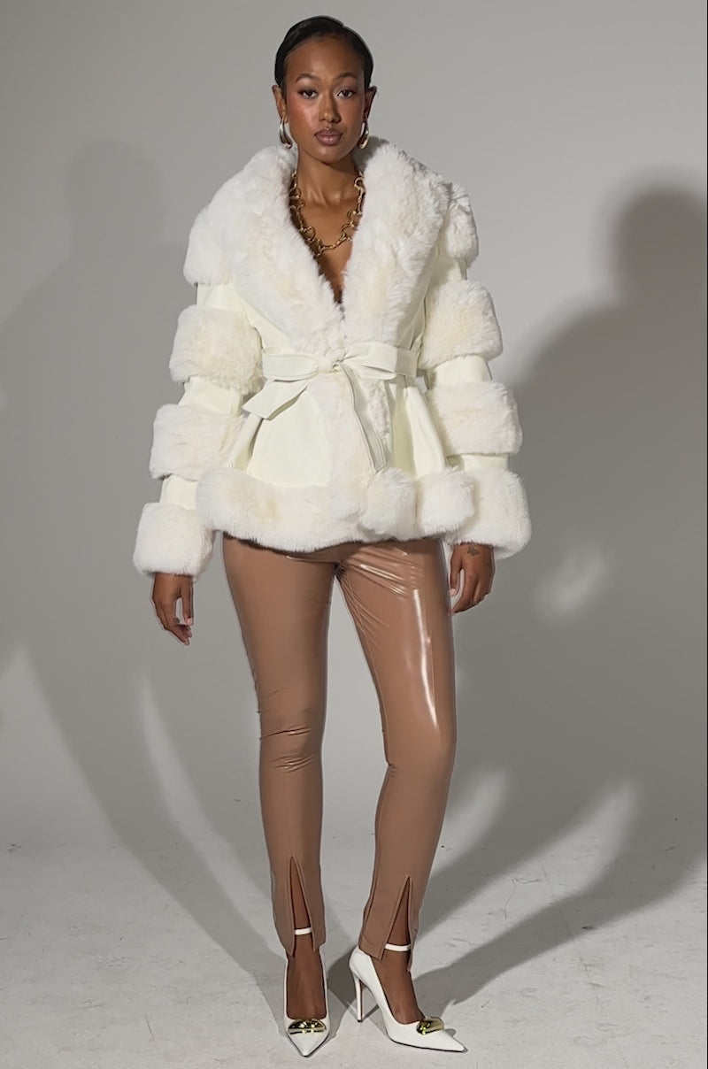A video of a woman wearing GET GROOVY FAUX LEATHER SKINNY LEGGING with a fluffy white faux fur jacket, satin belt, white pointed heels, and gold jewelry, standing against a plain light background.