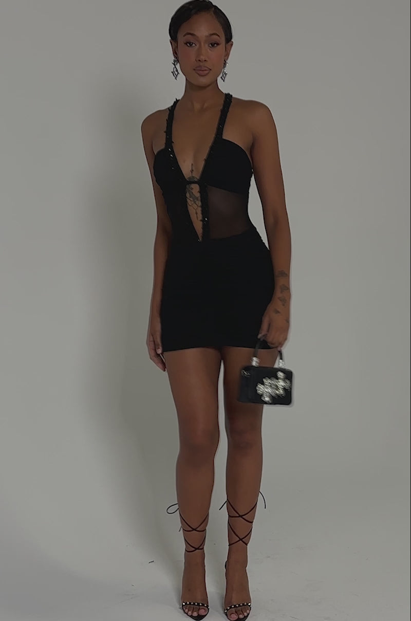 Video of woman modeling the SHAKE IT GIRL MINI DRESS, a fitted, ruched mesh mini dress featuring a deep plunging neckline and sheer panel. She has short hair, visible tattoos on her arm and hand, and poses against a plain background.