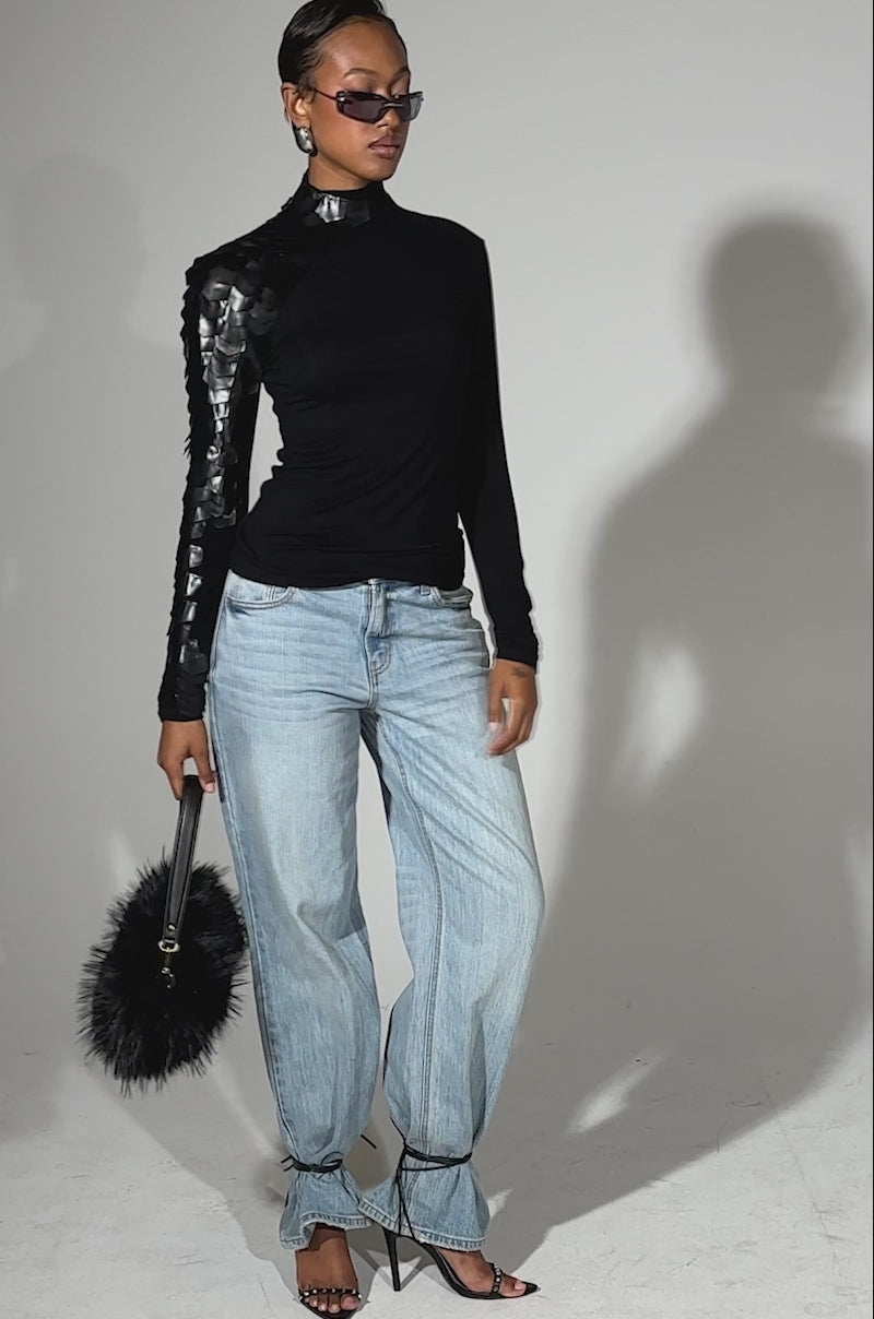 A video of a woman wearing the NASTASIA SCALE TOP—a fitted black turtleneck with geometric faux leather cutouts on one shoulder—paired with light blue high-waisted jeans, strappy black heels, sunglasses, and a fluffy black handbag.