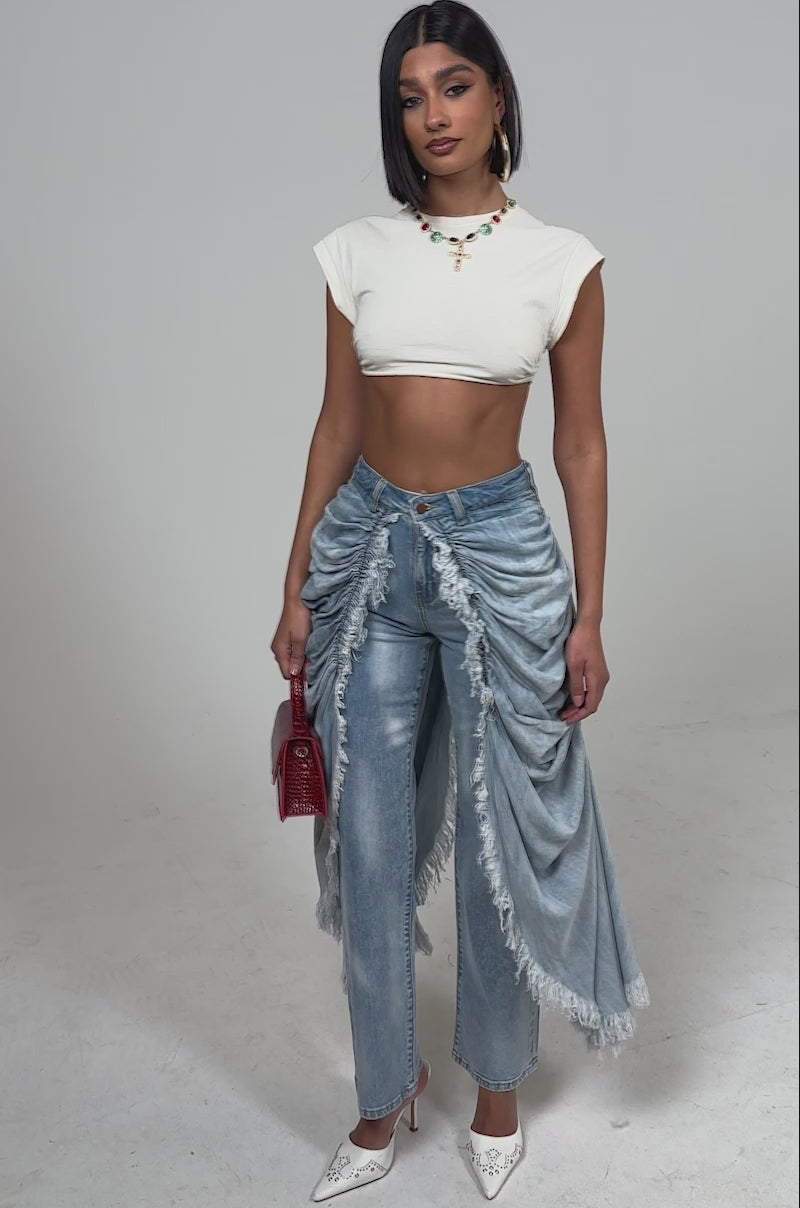 A video of a woman in the ALL OVER THE PLACE DENIM PANT—high-rise, light-wash jeans with dramatic, fringed front drapes—paired with a white crop top, red handbag, white heels, and a necklace.
