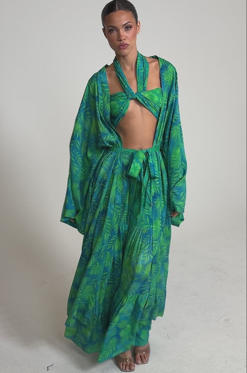 Video of woman modeling the INTO THE GARDEN DUSTER IN GREEN, a long, kimono-style dress with wide sleeves and a sash belt, showcasing its vibrant tropical print against a light gray background.