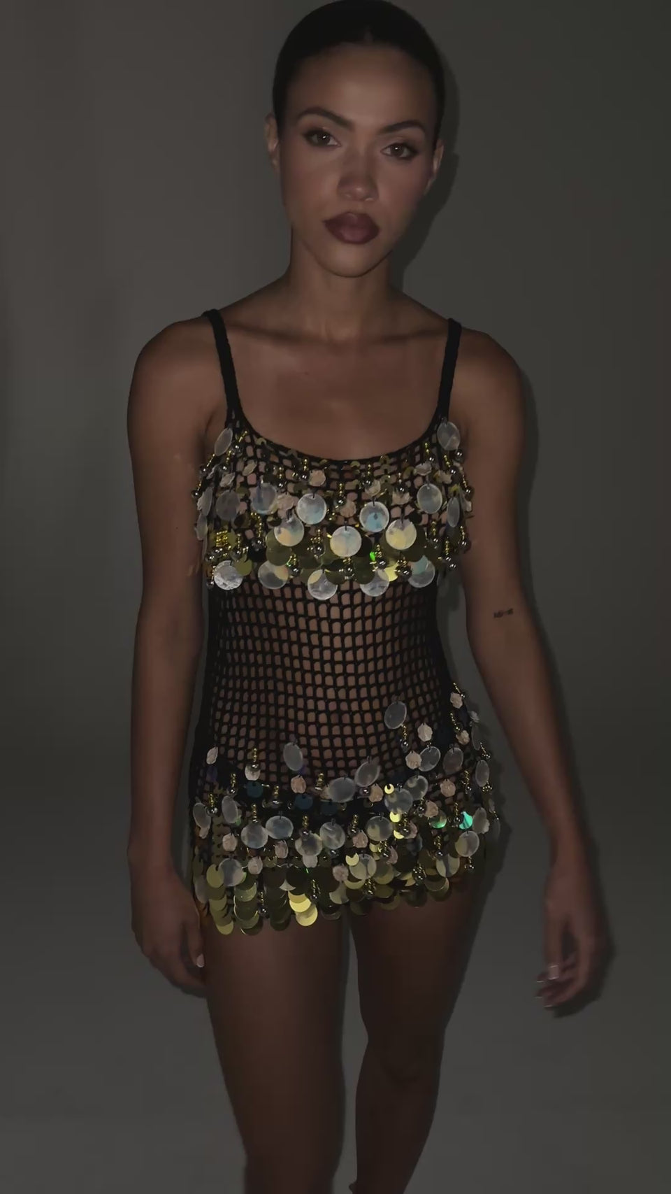 A video of a model wearing RESORT LIVING SEQUIN CROCHET MINI DRESS.  Featuring an open-knit crochet base, a scoop neckline, delicate shoulder straps, and an assortment of gold metallic sequins, beaded embellishments, and seashell accents lining the bust and hem.