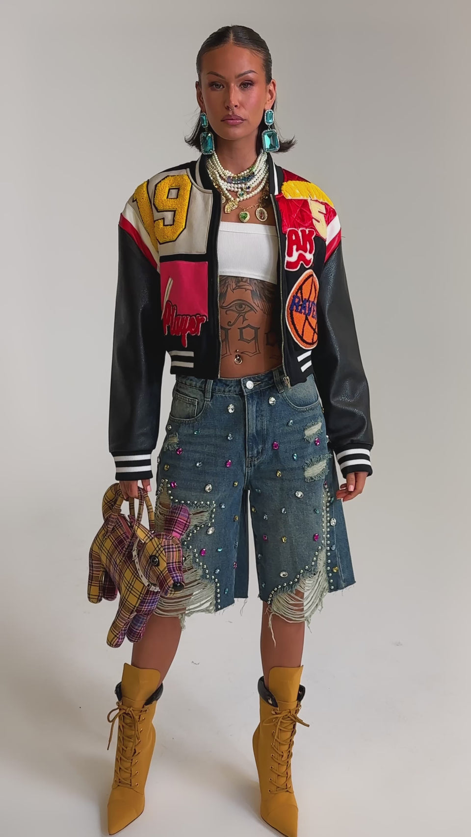 A video of a woman in the NEW YORK NEW YORK VARSITY BOMBER, paired with a white crop top, ripped decorated denim shorts, tan lace-up boots, statement earrings, and holding a patchwork stuffed animal.