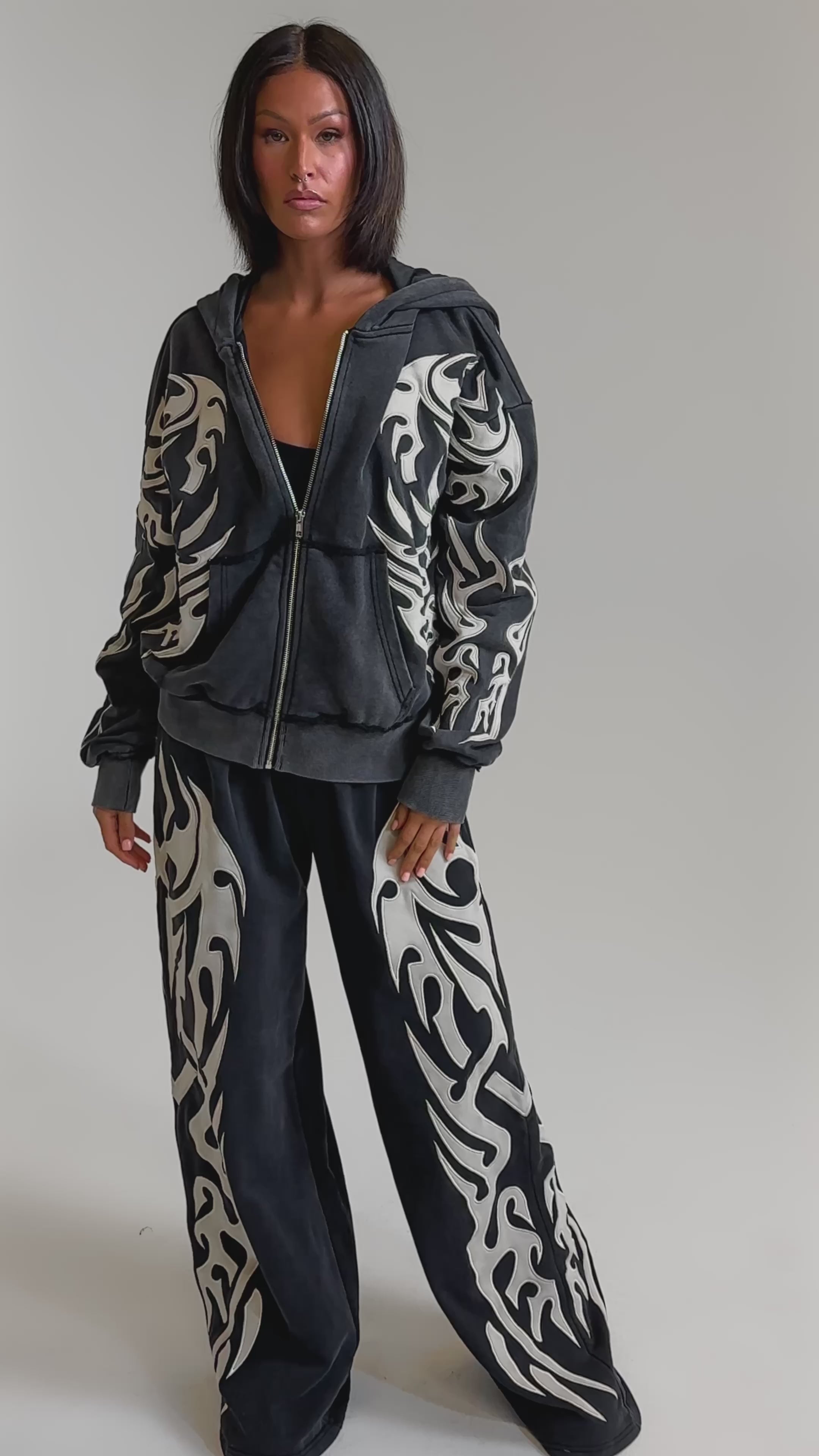 A video of a woman in the ON FIRE ZIP UP SWEATSHIRT—an oversized black velour tracksuit with bold white tribal patterns and an embroidered flame motif. 