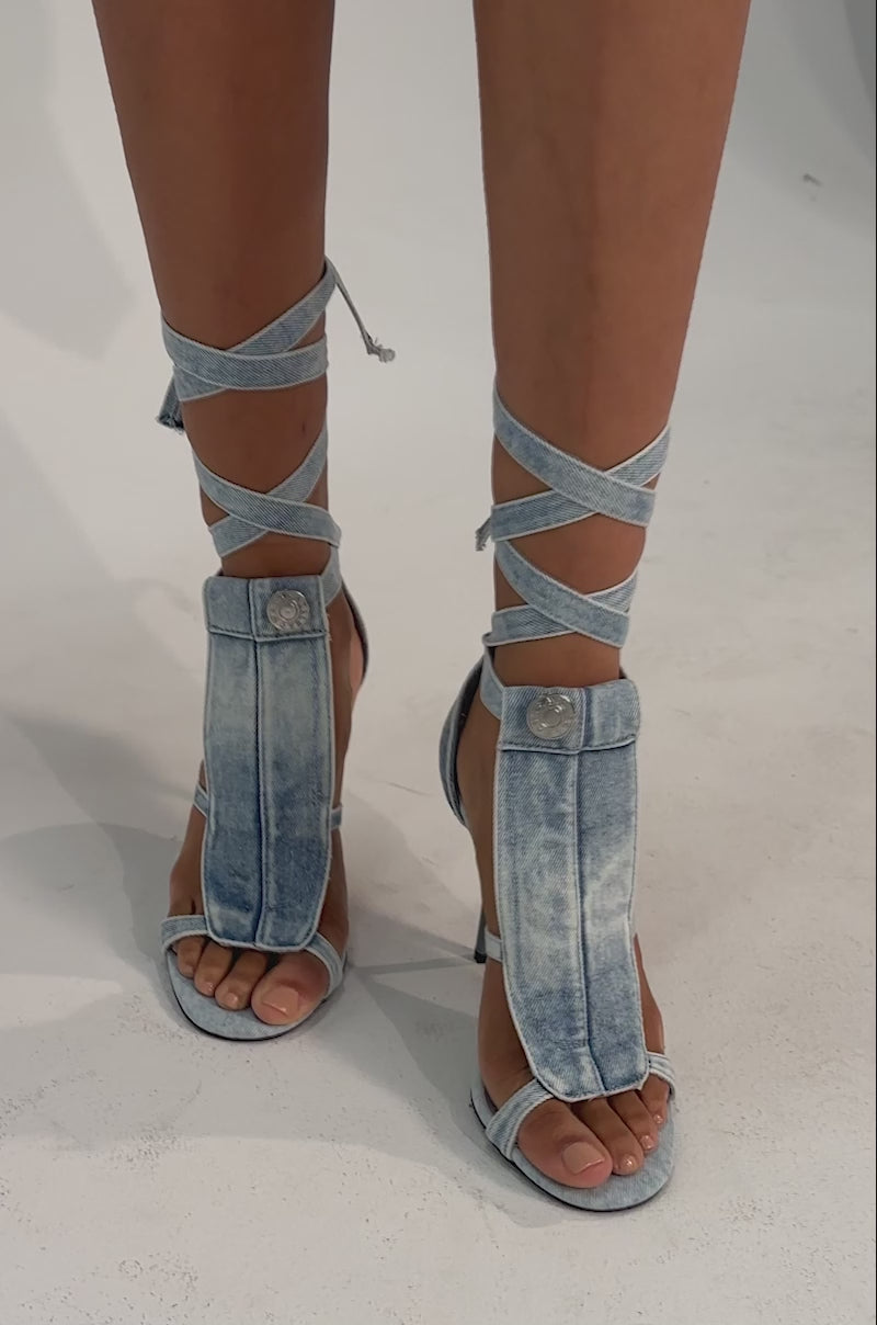 A video of a woman wearing the AKIRA GOLD NORMA JEAN DENIM STRAPPY STILETO SANDAL in light blue, featuring open toes and wrap-around ankle straps.