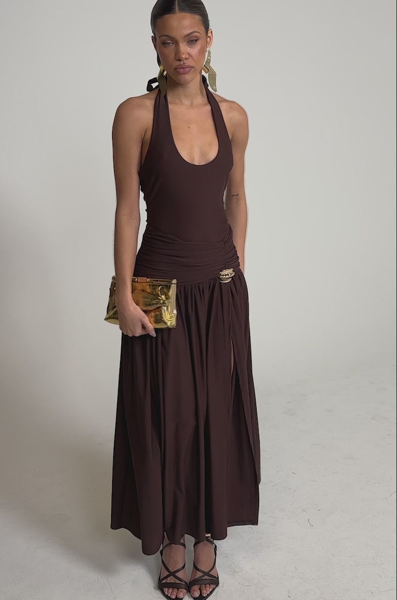 A video of a woman wearing the PERFECT RUCHED MAXI SKIRT in chocolate brown with a high slit, paired with a matching halter top, black strappy sandals, large gold earrings, and a gold textured clutch against a plain light background.
