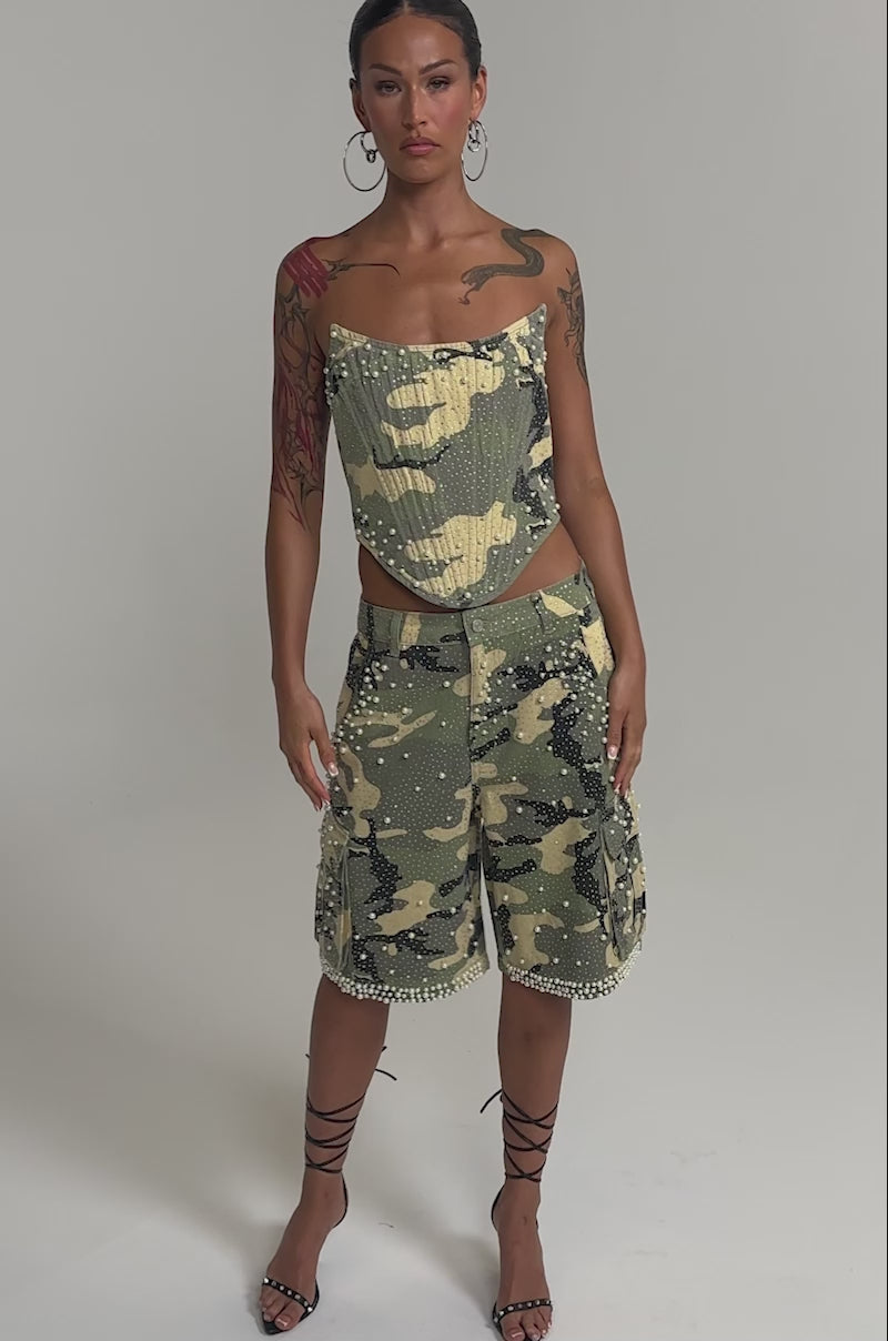 Video of woman modeling the EMBELLISHED CAMO CORSET, a strapless pearl rhinestone camo top paired with matching pants. She accessorizes with hoop earrings, showcases visible tattoos, and stands against a plain background.