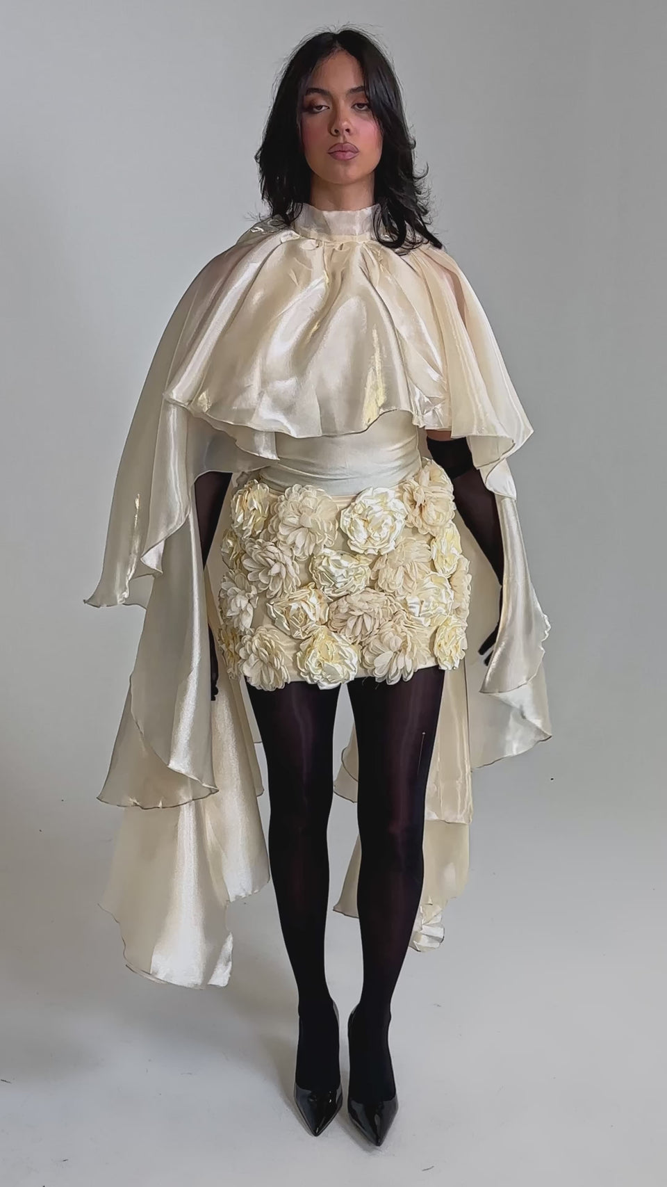 A video of a woman wearing the STAR OF THE NIGHT TOP, a high-neck ivory organza cape with dramatic ruffles, styled with a textured floral skirt, black sheer tights, gloves, and pointed black heels.
