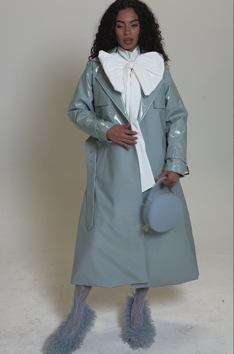 Video of woman modeling the LOCKED AND LOADED PATENT TRENCH in shiny light grey, styled with a white blouse, patterned tights, fluffy light blue heels, and a round light blue handbag against a plain background.