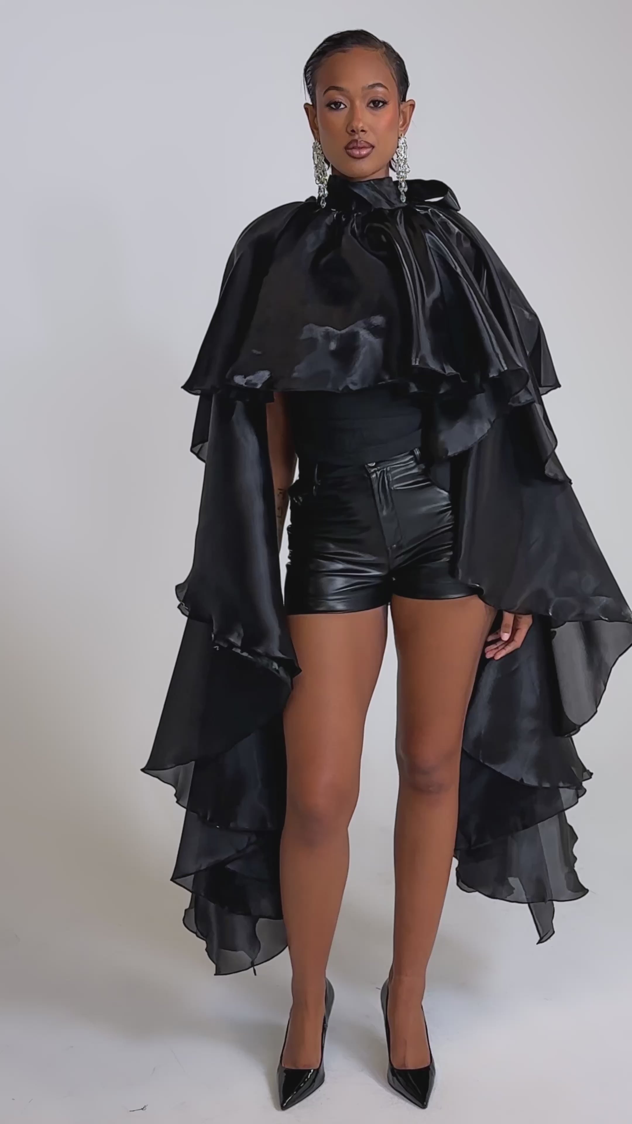 A video of a woman wearing the COOLEST ON THE BLOCK BLOUSE and leather shorts. Black heels and slicked-back hair complete her bold, modern look.