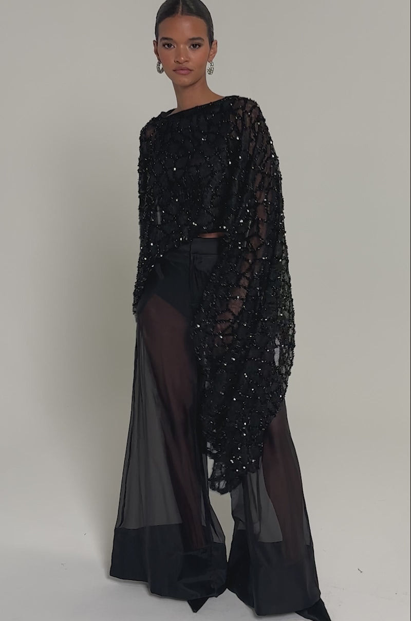  Video of woman modeling the SHINE AWAY MESH BLOUSE, featuring sheer mesh fabric, batwing sleeves, and sequin details, paired with wide-leg pants. She stands confidently against a plain light background.