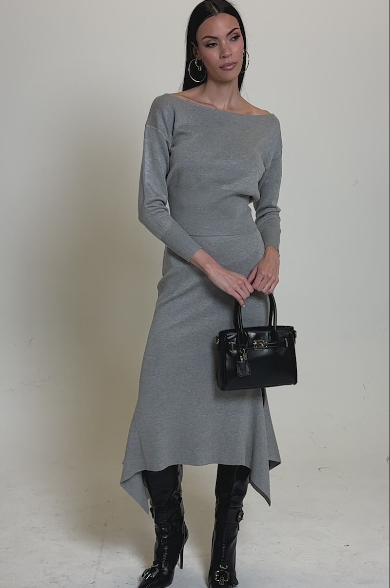 A video of a woman wearing the AKIRA Label LEXI OFF THE SHOULDER SWEATER, styled as a fitted gray dress with an asymmetrical hem, tall black boots, hoop earrings, and carrying a black handbag in her left hand.