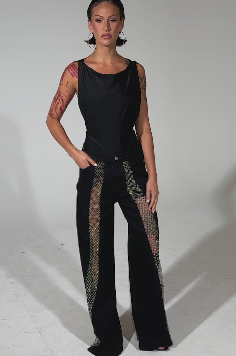 A video of a woman with slicked-back hair posing confidently against a plain, light background, wearing SOMETHING DIFFERENT RELAXED DENIM WITH RHINESTONE MESH and a sleeveless black top, one hand casually in her pocket.