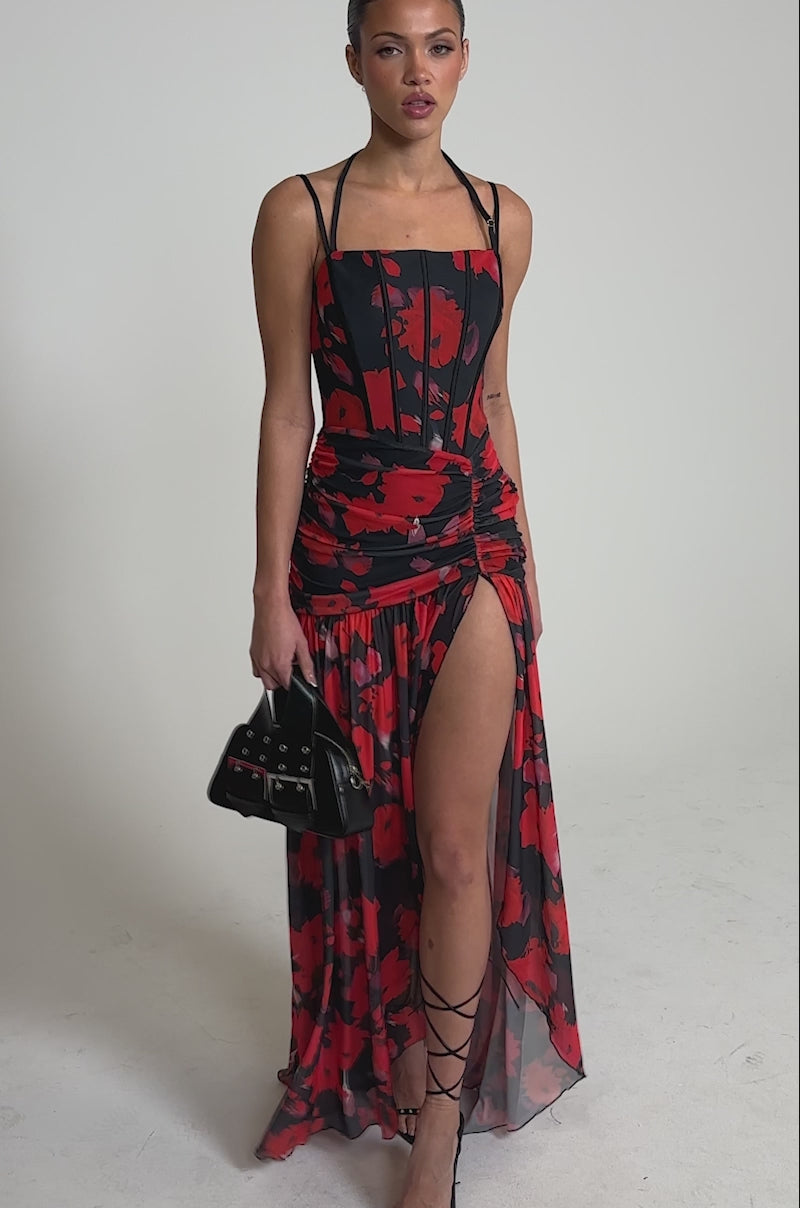 Video of woman modeling the ROSE GARDEN MESH MAXI DRESS, featuring a red and black abstract floral print, spaghetti straps, ruched details, a high slit, and corset-style bodice, styled with strappy heels.
