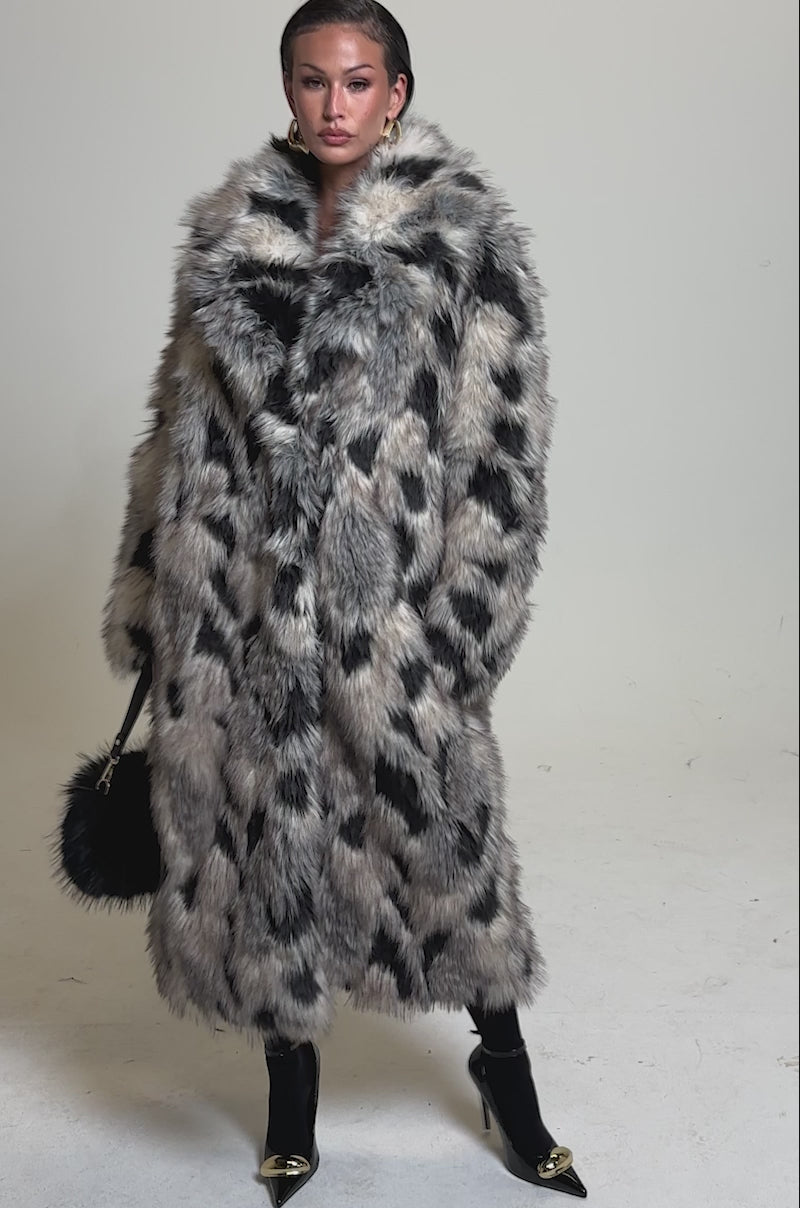 A video of a model wearing the NEW AUSTIN FAUX FUR COAT in an oversized, black and white spotted design, this person exudes vintage luxury paired with a black furry handbag, gold-accented black heels, slicked-back hair, and bold earrings.