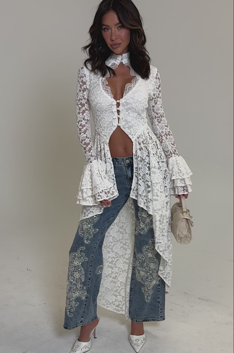 Video of woman modeling the BILLIONAIRE BELLE RHINESTONE AND PEARL JEANS with intricate white lace and sequin floral embroidery, paired with a white lace ruffled-sleeve top and white pointed-toe heels.