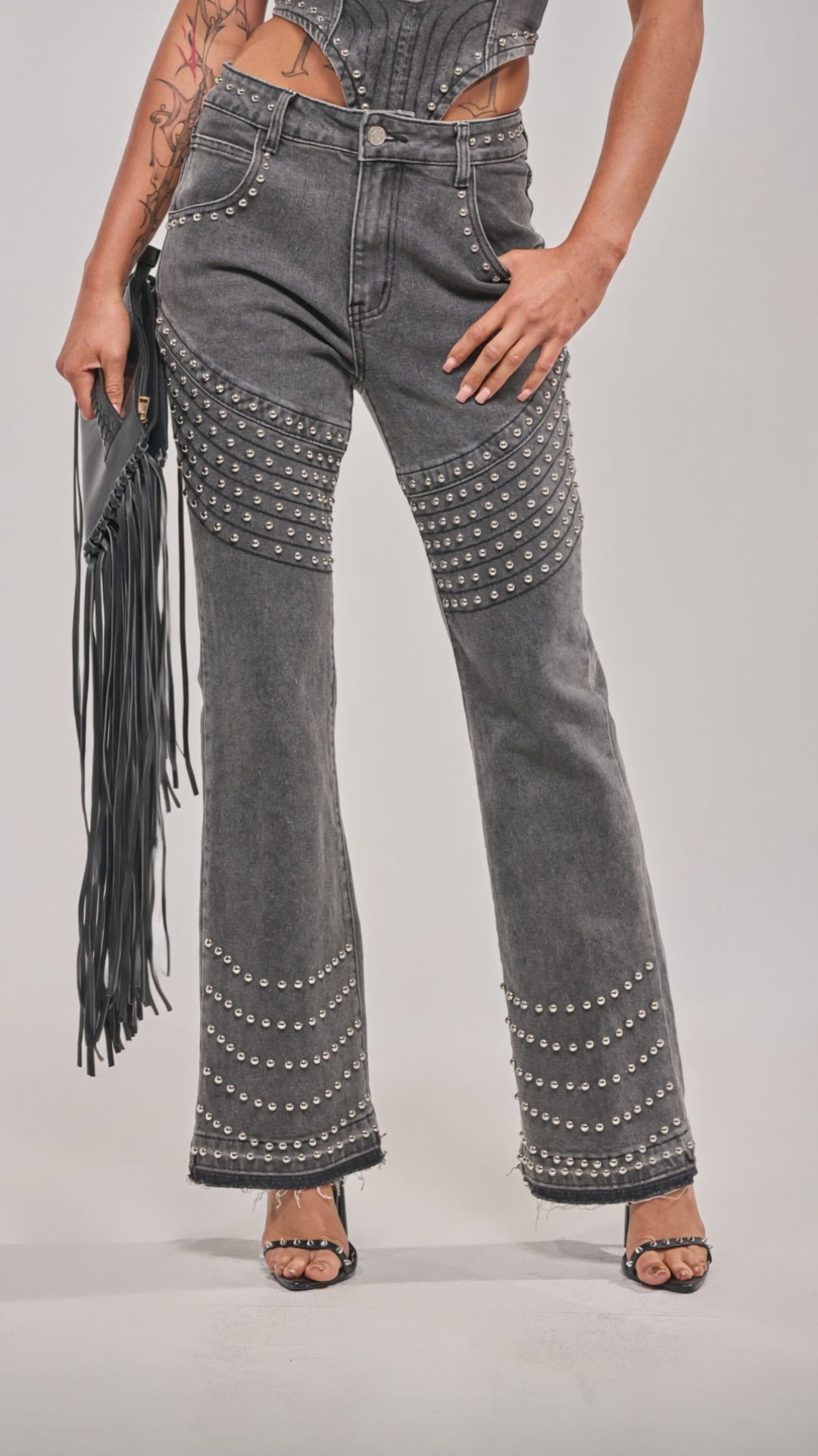 Video of woman modeling the FEELING THE VIBES FLARE DENIM—a strapless, structured grey top and matching flare jeans with dome stud accents. She accessorizes with a black fringe clutch, heels, and displays tattoos against a light grey background.