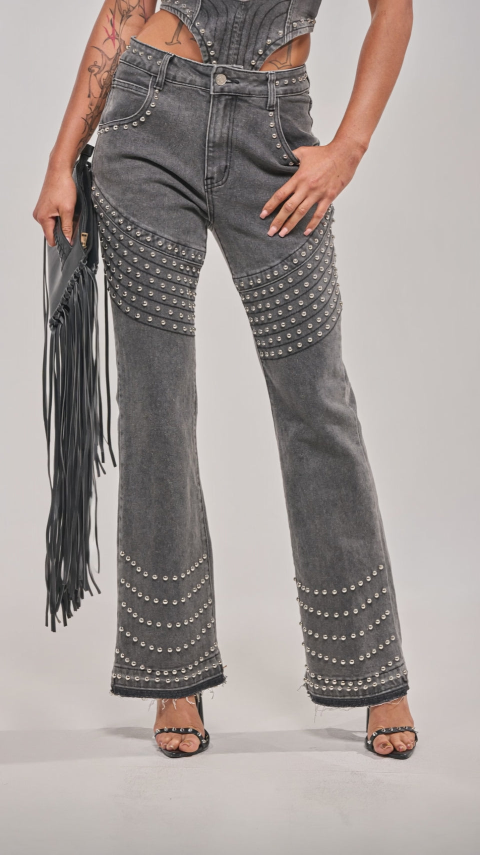 Video of woman modeling the FEELING THE VIBES FLARE DENIM—a strapless, structured grey top and matching flare jeans with dome stud accents. She accessorizes with a black fringe clutch, heels, and displays tattoos against a light grey background.