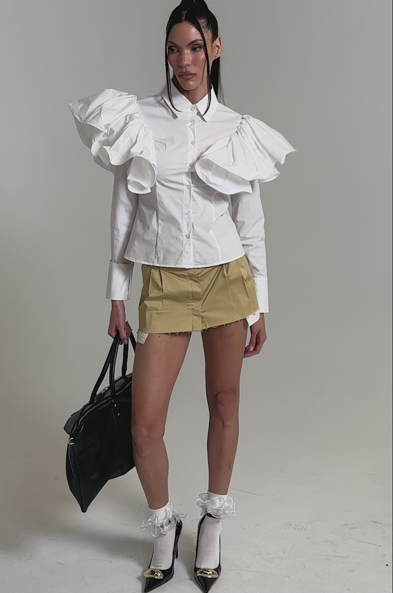 A video of a woman wearing the NO BIG DEAL BUTTON DOWN, a white blouse with large ruffled sleeves, paired with a short tan skirt, sheer socks with white ruffles, black heels, and carrying a large black handbag.