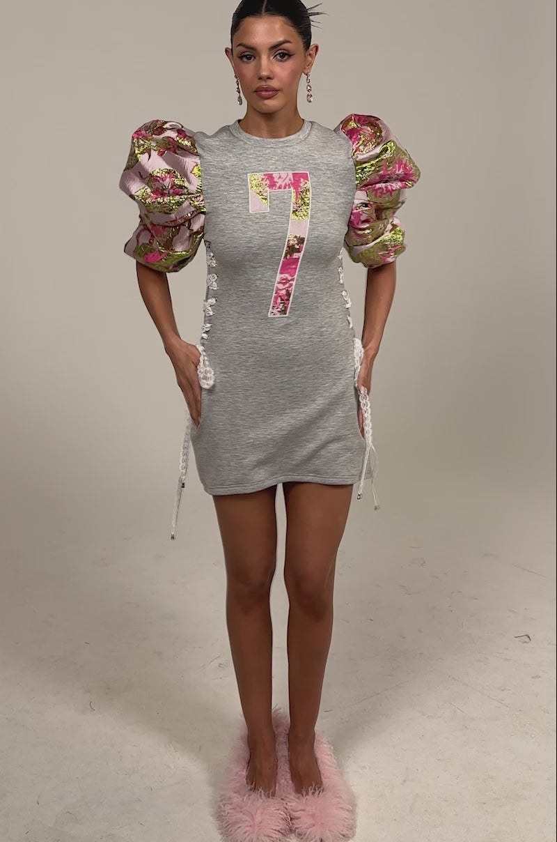 Video of woman modeling the GAME CHANGER BROCADE LACE UP MINI DRESS, a short grey dress featuring a bold pink floral number 7 and puffed floral-pattern sleeves