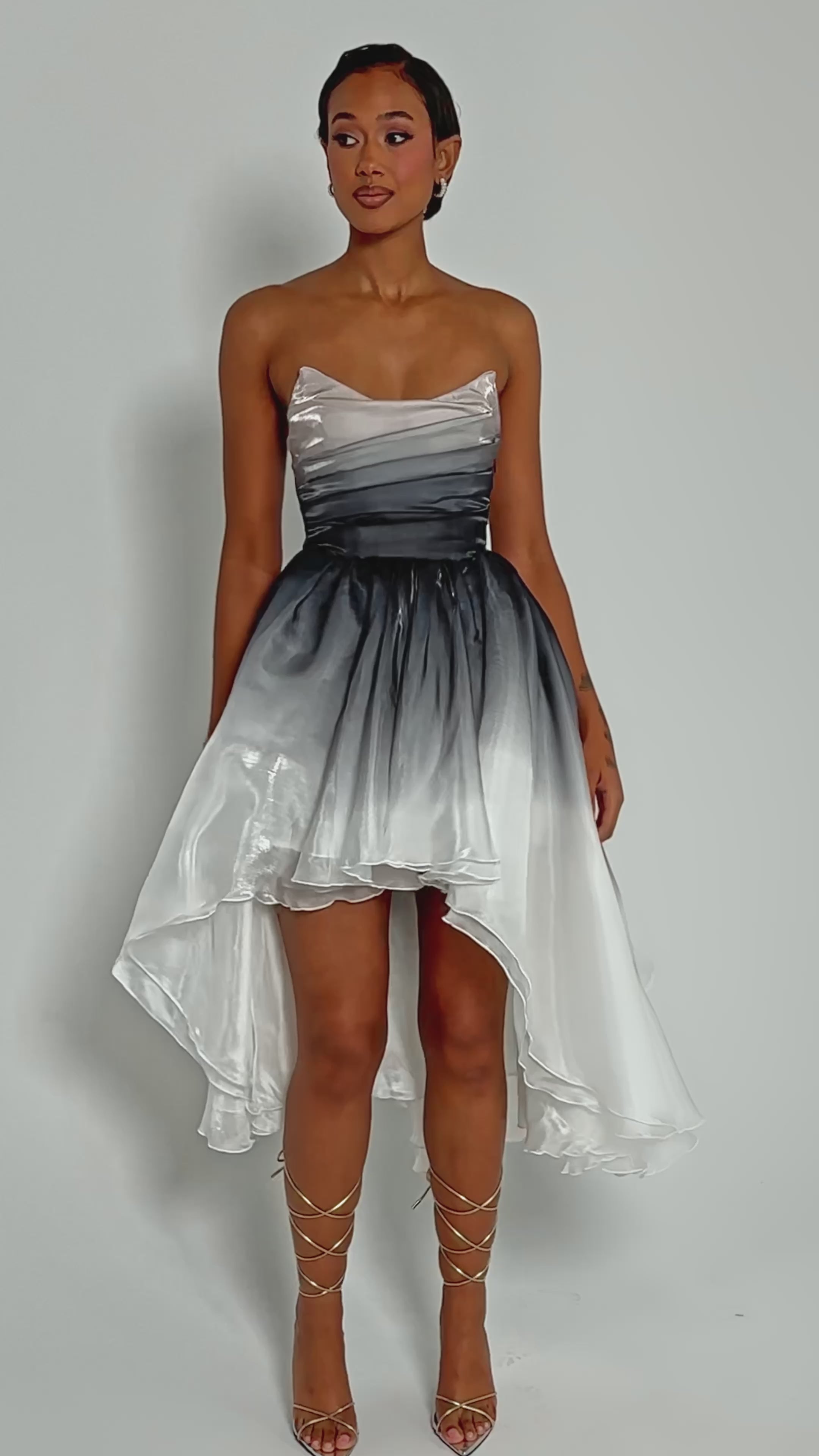 Video of woman modeling the DANCE WITH ME HIGH LOW OMBRE DRESS, featuring a strapless fitted bodice and a high-low ombre design that fades from white to dark gray and back to white, standing against a plain background.