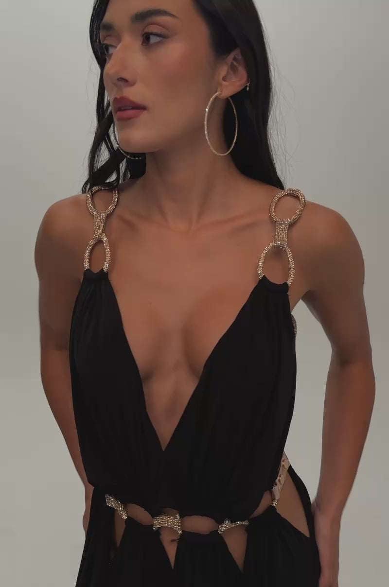 Video of a woman modeling the SITTING PRETTY PAVE CHAIN LINK JUMPSUIT DRESS. The sleek black jumpsuit featuring a dramatic plunge V-neckline, waist cut-outs adorned with metal rings and rose gold rhinestone chain accents, and elegantly flowing wide-leg pants.